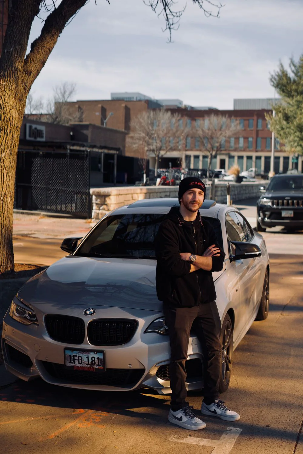 A man in black standing in front of a silver BMW sedan downtown.